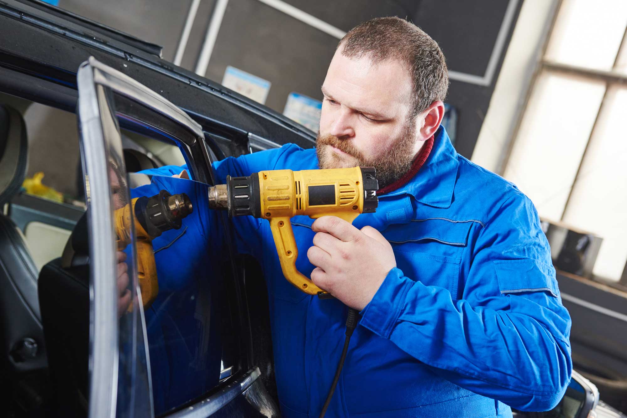 Auto Glass Repair Services In this image, a person is wearing a blue work uniform and using a yellow power drill on the window of a car. The car, which is dark-colored, appears to be inside a workshop. In the background, there are organized shelves or racks, suggesting a professional setting. The worker seems to be focused on installing or repairing something related to the car’s window.