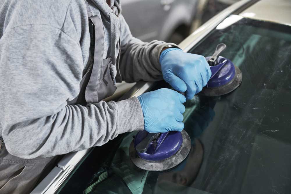n this image, a person is wearing a grey hoodie and blue gloves, diligently working on a car’s windshield. The individual is using two suction cup tools attached to the car’s windshield, likely for repair or replacement. The focus of the image is on the person’s hands and tools, illustrating the technical aspect of the work being performed. The car has a dark-colored exterior, but specific details are not visible due to the angle of capture. The background appears to be a workshop or garage setting, although it isn’t clearly visible.