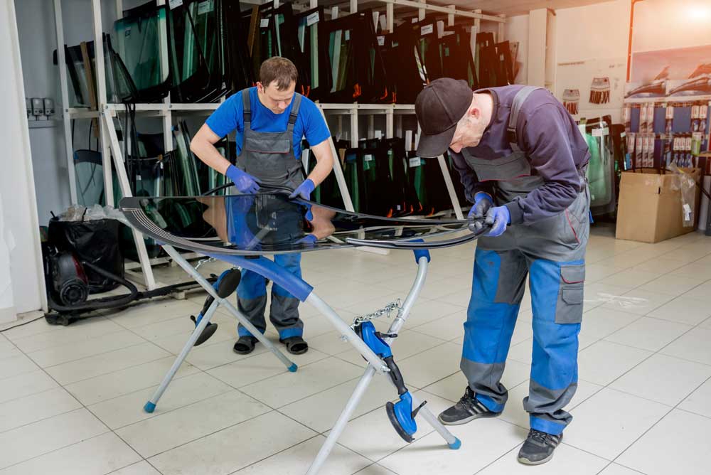 Trusted Auto Glass Shop In this image, two individuals are working together to install or repair a car windshield. They are in a workshop environment, surrounded by shelves holding various pieces of glass, likely replacements for different vehicle models. Both workers are dressed in dark blue work attire with gloves to ensure safety and precision in their task. The worker on the right is bending over slightly, focusing on aligning or fitting the glass properly. The setting is a well-organized workshop with shelves lined with various sizes and shapes of glass panels in the background. A specialized stand with adjustable arms supports the windshield, aiding the workers in their task. The floor is clean and reflects the professional environment of the workshop