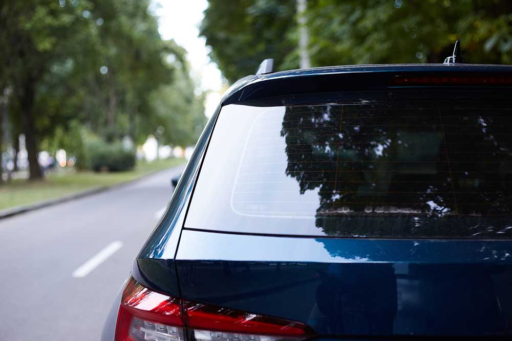 Back Window Replacement in Baltimore This image captures the rear part of a dark blue car parked on the side of a road, surrounded by lush greenery. The car’s rear window is slightly tinted, reflecting the trees and sky. The vehicle’s brake light is visible, and it appears to be in pristine condition.