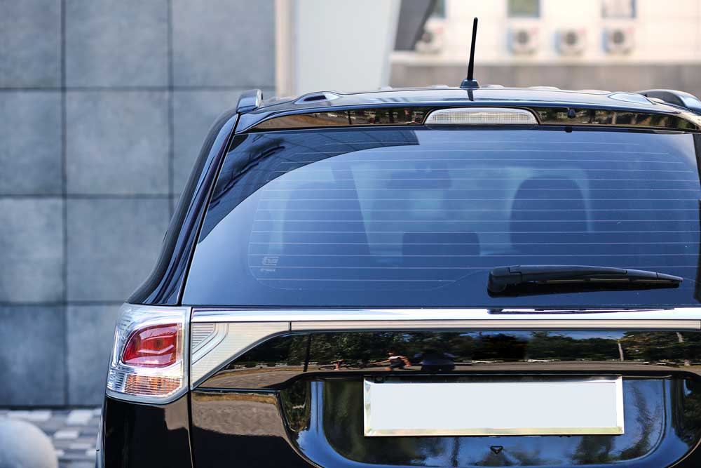 High-quality Back window replacement This image captures the rear view of a dark-colored car parked in front of a grey, stone wall. The car’s glossy finish is evident, reflecting the surrounding environment. The rear windshield is large and slightly curved with horizontal lines running across it. A windshield wiper is visible at the bottom of the rear windshield. The taillights are red and white, with a modern design that aligns with the shape of the car.