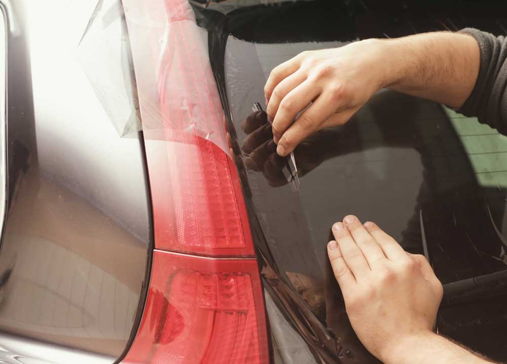 Reliable Back Window Replacement This image captures a moment where a person is applying tint to the window of a car. The individual’s hands are focused on smoothing out the film, ensuring it adheres properly to the glass. The car’s red tail light is visible, and it appears that the window tinting process is being done with precision and care
