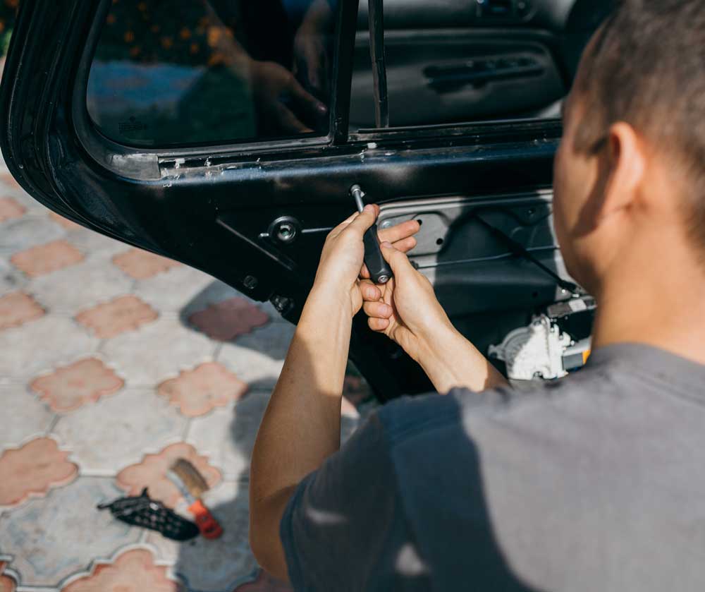 Passenger Window Replacement Process In this image, a person is working on a car door that is open; their face is blurred out. The individual appears to be using a screwdriver or similar tool to work on the internal mechanisms of the car door. Various tools and parts are scattered on the ground next to where they’re working. The setting seems to be outdoors as natural light illuminates the scene and patterned paving stones are visible underfoot. The interior side of the car door is exposed, showing its mechanical components.