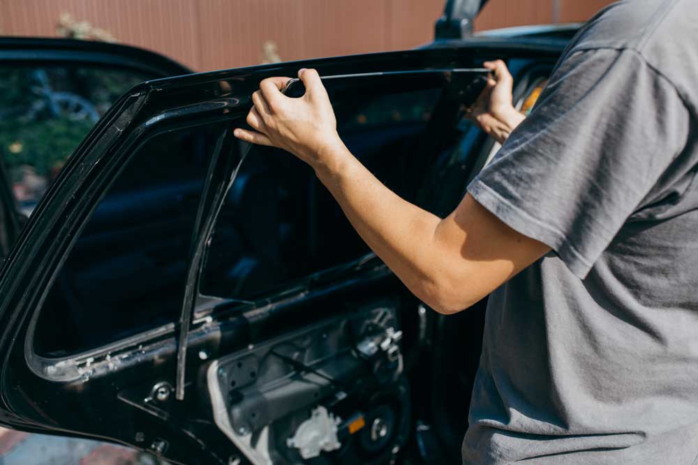 Passenger Window Replacement In the image, a person wearing grey clothing is working on a black car. The individual’s hands are focused on the upper part of an open car door, which has had its interior panel removed. This exposes wires and mechanical components within the door. The scene appears to be daytime, with sunlight illuminating part of the area.