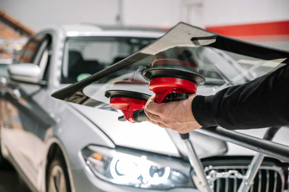 Windshield Repair Solutions In the image, a person is using a specialized tool with red suction cups to install or repair a windshield on a silver car. The car is parked indoors, possibly within a garage or workshop, illuminated by bright lighting. The individual is wearing dark clothing and appears to be professional, suggesting that this might be an auto repair shop where vehicles are serviced. The process seems precise and deliberate, emphasizing the importance of proper windshield installation for safety and functionality.