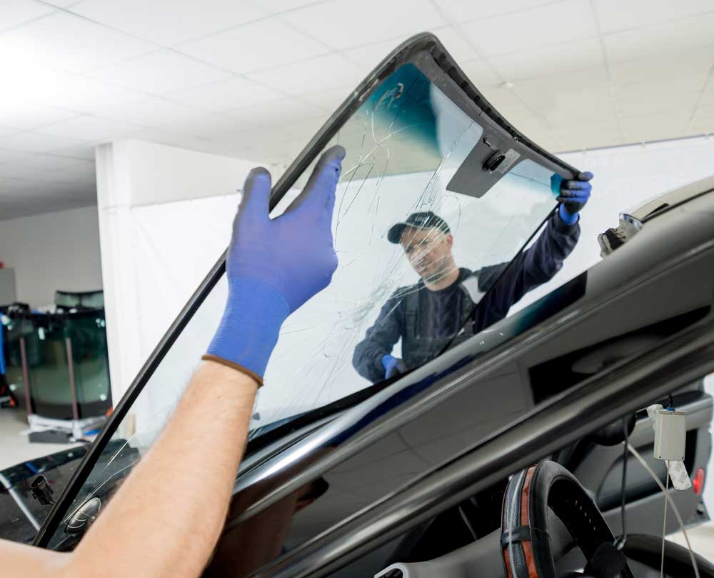 Windshield Repair in Baltimore In the image, a person wearing blue gloves is carefully installing or replacing a cracked or damaged car windshield. The individual is positioning the glass onto the exposed area of the car, which awaits the new installation. The setting appears to be a professional garage or workshop, with other replacement windshields stored vertically against the wall in the background. Tools and equipment are visible, indicating an ongoing repair or replacement job