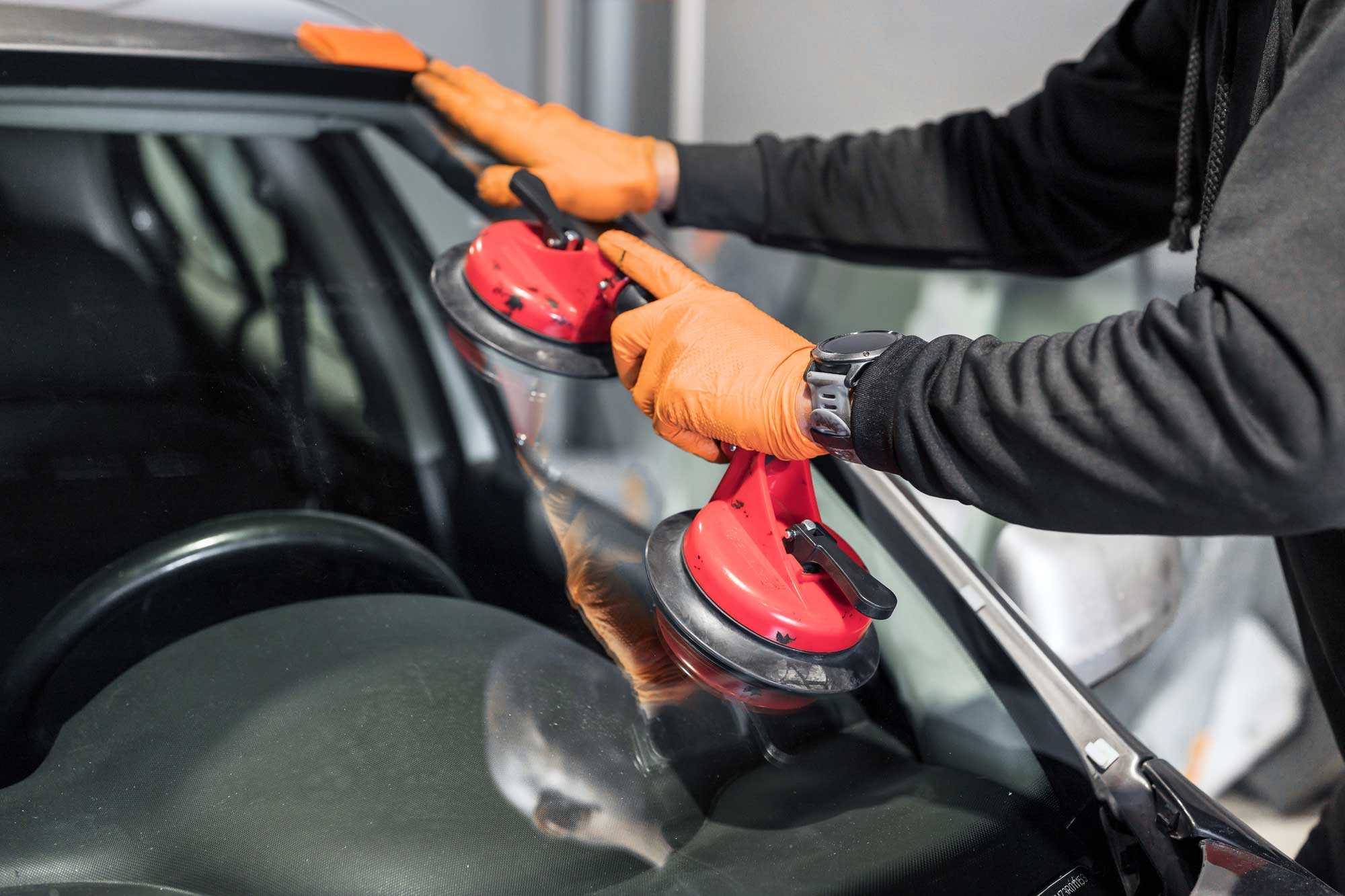 This image depicts a person wearing orange gloves and a dark long-sleeve shirt, diligently working on a car’s windshield. The individual is utilizing two red suction cup tools to either hold or install the windshield. The car’s hood is open, revealing the engine bay covered by a protective cloth. The setting suggests an indoor environment, likely a professional garage or workshop. 
