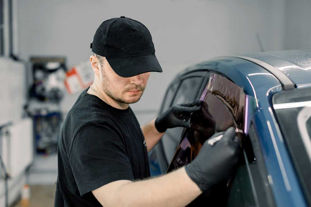 In this image, a person is seen applying tint to the window of a car inside what appears to be a workshop. The individual is wearing black gloves and is in the process of smoothing out the tinted film on the car’s window to ensure it adheres properly and is free of bubbles or wrinkles. The car has a shiny, metallic finish, and various items can be seen in the background, indicating that this setting is likely a professional workspace for vehicle modifications or repairs. 