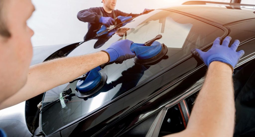 two workers in blue gloves installing a windshield on a black car using suction cups, with sunlight reflecting off the glass in a professional setting.