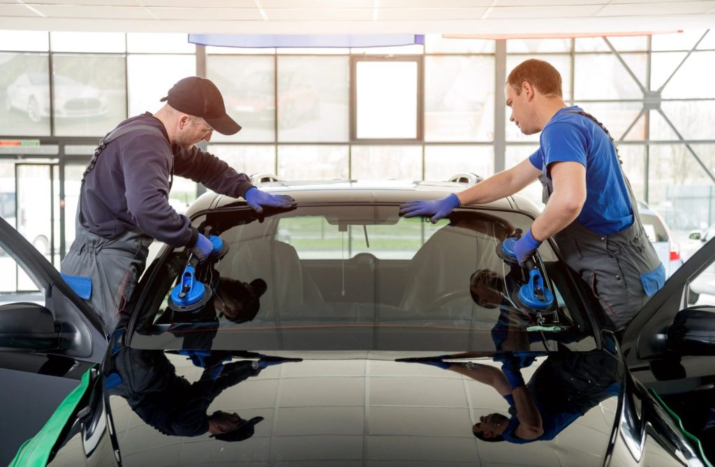 Professional workers from auto glass shop Baltimore installing new windshield on black vehicle using blue suction handles in bright modern service bay.