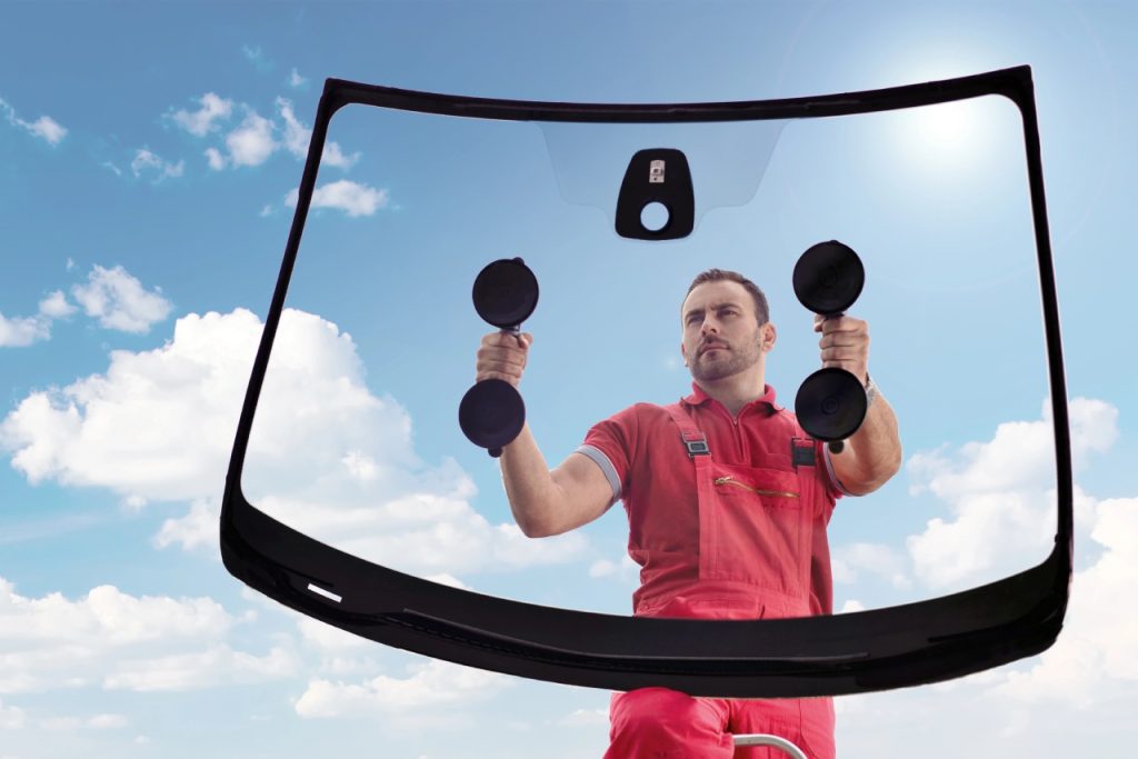 Expert at a leading auto glass company Baltimore holds a new windshield with suction cups against a clear blue sky background.